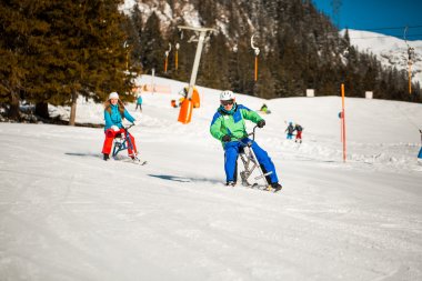 Zwei Personen fahren auf Skiern die Piste hinunter. Strahlender Sonnenschein und schneebedeckte Berge bieten eine schöne Kulisse.