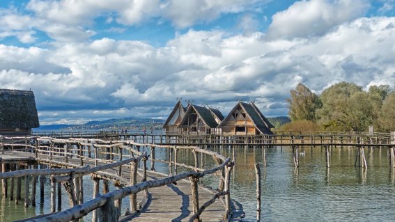 Holzhäuser stehen am Wasser. Ein Holzsteig führt zu den Häusern. Der Himmel ist bewölkt und der Tag ist hell.