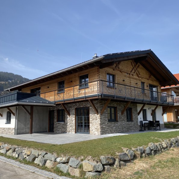 Das Haus steht in den Bergen. Es hat einen Balkon und ist aus Holz. Die Sonne scheint auf die Fassade.