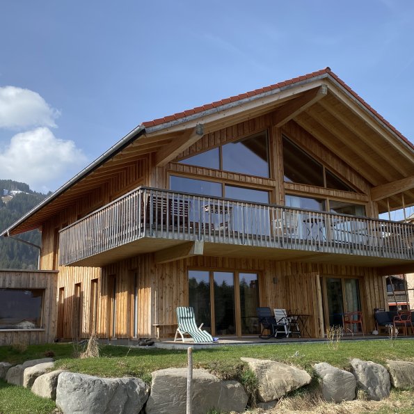 Ein Holzhaus steht in den Bergen. Menschen sitzen auf der Terrasse. Die Sonne scheint auf das Haus.