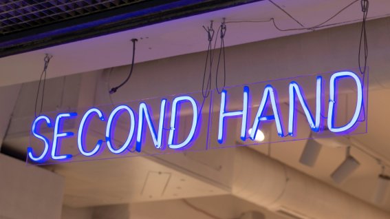 Ein Neonschild leuchtet mit der Aufschrift Second Hand in einem Raum. Das Licht strahlt in blauen Farben und zieht die Aufmerksamkeit an.