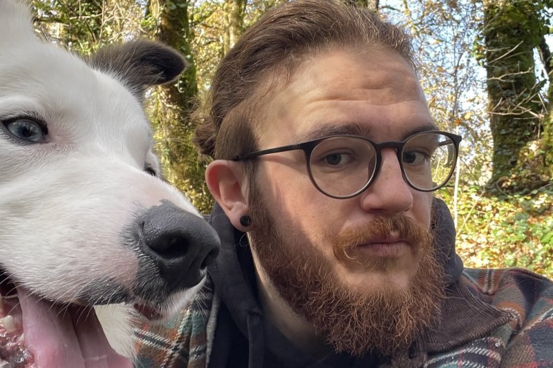 Ein Mann mit Bart posiert lächelnd neben einem Hund im Wald, umgeben von Bäumen und natürlichem Licht.