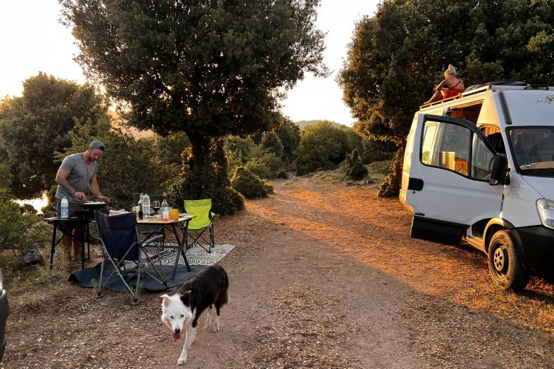 Ein Mann grillt während des Campingurlaubs am Fluss. Sein Hund läuft um den Wohnmobilplatz. Entspannung in schöner Umgebung.