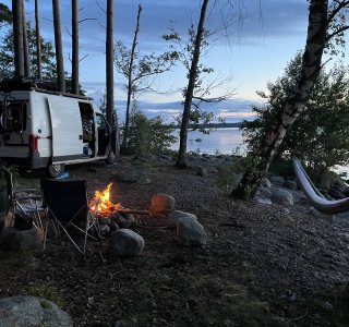 Ein Wohnmobil steht am Ufer eines Sees, während ein Lagerfeuer brennt und ein Hängematte zwischen Bäumen schwingt.