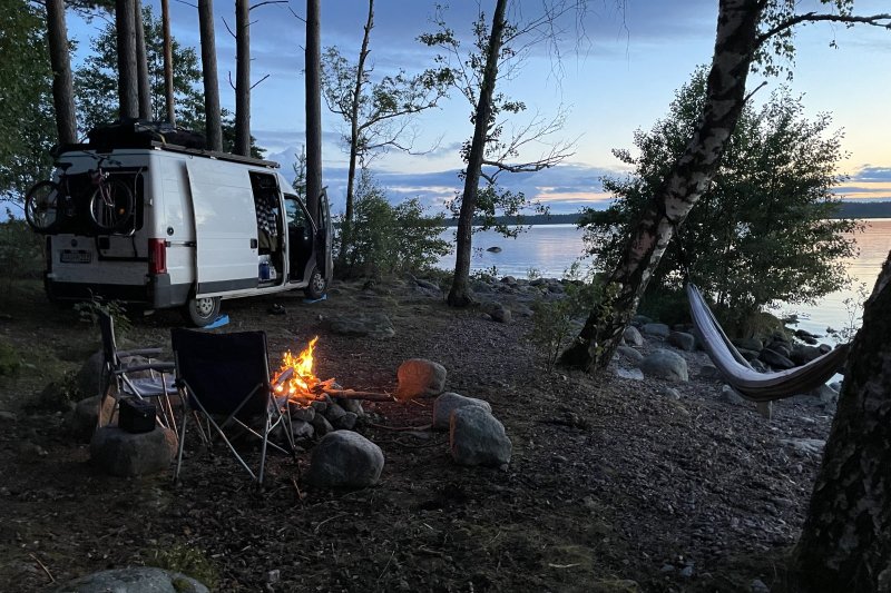 Ein Camper-Van parkt zwischen Kiefern am Seeufer, davor knistert ein Lagerfeuer, Campingstühle und eine Hängematte laden zum Verweilen ein. Freiheit und Naturerlebnis, die den Pura-Vida-Gedanken prägen.