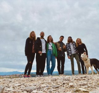 Eine Gruppe von sechs Freunden posiert fröhlich auf einer Steinlandschaft mit einem Hund im Hintergrund bei bewölktem Wetter.