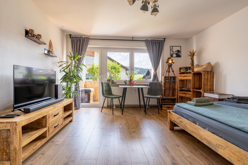 Der Raum hat ein Bett, einen Tisch und einen Fernseher. Das Fenster bietet einen Blick auf die Natur draußen.