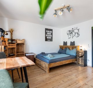 In einem Raum stehen ein Holzbetten und ein Tisch. Es gibt Pflanzen und Licht. Es ist ein heller Raum am Tag.