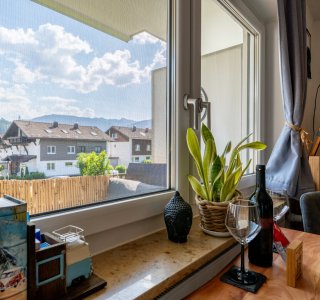 Durch ein großes Fenster ist eine Stadt zu sehen. Draußen stehen Häuser und Bäume. Es ist sonnig und blau.