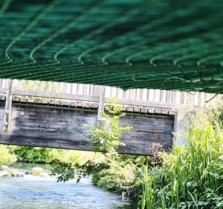 Eine Brücke steht über einem Fluss. Grüne Pflanzen wachsen am Ufer. Die Sonne scheint und das Wasser fließt.