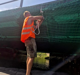 Ein Mann arbeitet an einer Brücke und bindet ein Seil fest. Es ist sonnig und der Ort ist an einem Gewässer.