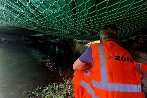 Der Mann sitzt am Ufer und schaut ins Wasser. Er trägt eine leuchtende Weste. Es ist tagsüber und die Umgebung ist grün.