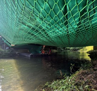 Menschen arbeiten unter einer Brücke. Sie reparieren die Struktur. Der Fluss fließt ruhig darunter. Es ist hell am Tag.