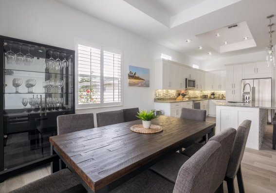 In einem hellen Raum stehen ein großer Tisch und eine moderne Küche. Der Raum hat Fenster und eine angenehme Atmosphäre.