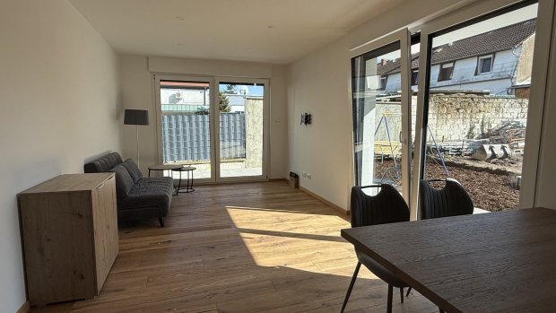 Ein Wohnzimmer hat große Fenster und viel Licht. Es gibt eine Couch, einen Tisch und einen Blick auf den Garten draußen.