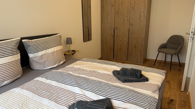 Im Raum steht ein großes Bett mit gestreiften Bettlaken. Es gibt einen Kleiderschrank und einen Stuhl in der Ecke.