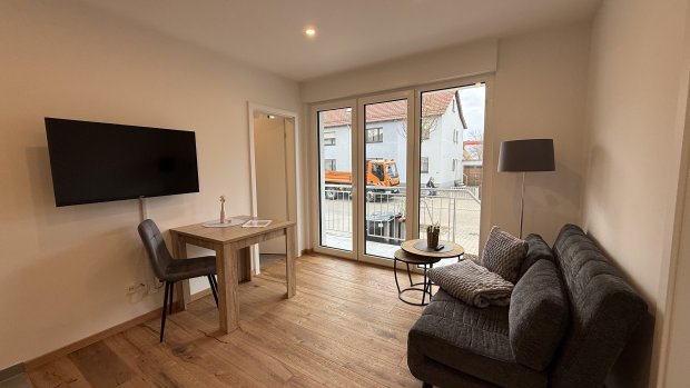 Im Raum sieht man ein Sofa, einen Tisch und einen Fernseher. Draußen sind Fahrzeuge und ein Hof zu sehen.