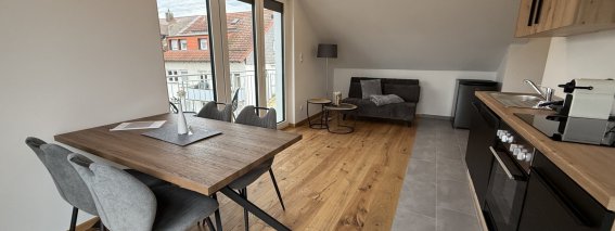 In einer Wohnung befinden sich ein Esstisch, Stühle und eine Küchenzeile. Der Raum hat große Fenster und viel Licht.