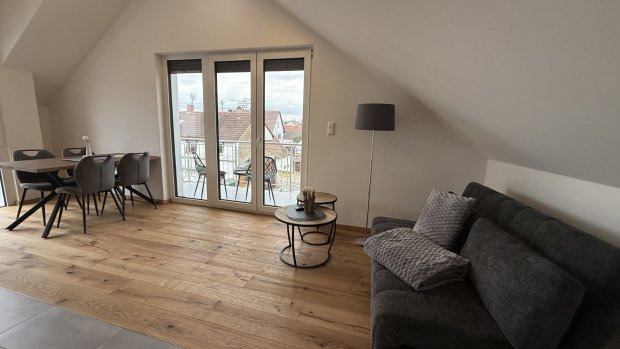 Ein Wohnzimmer mit einem Sofa und einem Tisch in einer Dachwohnung. Der Blick geht auf den Balkon und die Stadt.