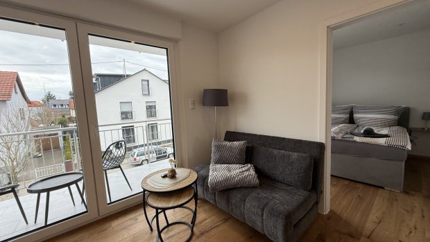 In einem modernen Raum stehen ein Sofa und ein Tisch. Die Tür führt zu einer Terrasse. Ein Bett ist im Hintergrund sichtbar.