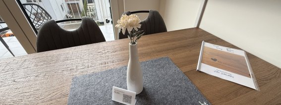 Der Tisch zeigt eine Vase mit einer Blume. Es gibt eine Tischunterlage und einen Informationszettel.