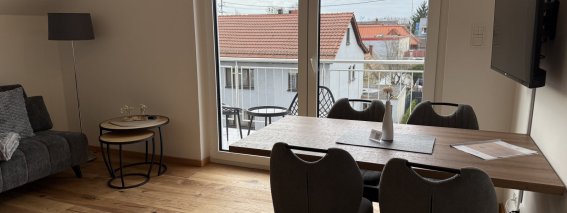 Das Wohnzimmer zeigt einen Tisch mit Stühlen und einen Fernseher. Der Blick geht nach draußen auf ein Gebäude.