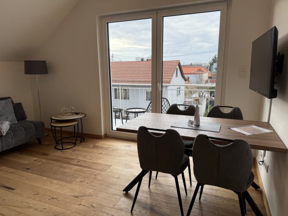 Das Wohnzimmer zeigt einen Tisch mit Stühlen und einen Fernseher. Der Blick geht nach draußen auf ein Gebäude.