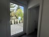 Durch ein großes Fenster sieht man einen Garten mit Bäumen und einer blauen Toilette. Der Himmel ist klar.