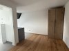 In einem kleinen, leeren Zimmer steht ein Schrank aus Holz. Der Boden ist aus Holz und die Wände sind hell gestrichen.