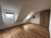 Ein leeres Zimmer mit schrägen Wänden und einem Fenster zeigt einen Holzfußboden. Die Wände sind weiß und ohne Möbel.