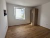 Der Raum hat einen Holzfußboden und ein Fenster, das Tageslicht hereinlässt. Es sind keine Möbel sichtbar.
