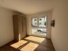 In einem hellen Zimmer steht ein Kleiderschrank neben einem Fenster. Draußen sind Fahrzeuge auf der Straße zu sehen.