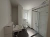 In einem neuen Badezimmer stehen eine Dusche, ein Waschbecken und eine Toilette. Der Raum ist hell und einfach gestaltet.