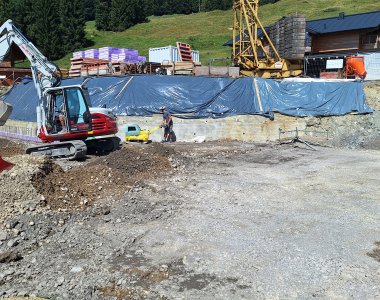 Auf einer Baustelle arbeiten Bagger und Bauarbeiter an einem Fundament in einer bergigen Umgebung unter klarem Himmel.