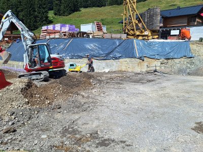 Auf einer Baustelle arbeiten Bagger und Bauarbeiter an einem Fundament in einer bergigen Umgebung unter klarem Himmel.
