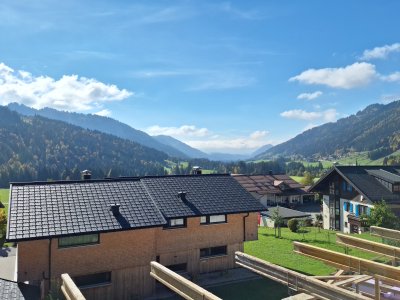 Die Landschaft im Herbst zeigt grüne Wiesen, Berge und Wolken. Die Häuser sind typisch für die Alpenregion.