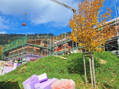 Auf der Baustelle wird an einem neuen Gebäude gearbeitet. Bäume zeigen herbstliche Farben unter blauem Himmel.