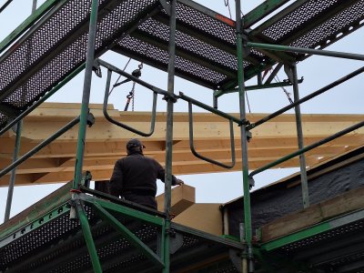 Ein Bauarbeiter arbeitet auf einem Gerüst und hebt Holzplatten in die Höhe. Es ist ein klarer Tag im Freien.
