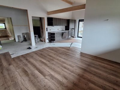 In der Wohnung wird der Boden verlegt. Die Küche und der Wohnbereich sind sichtbar. Es sind noch Arbeiten im Gange.