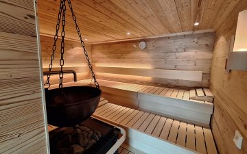 Finnische Sauna mit Aufgußautomat In der Sauna sitzen Personen auf Bänken aus Holz. Der Saunaofen steht in der Mitte. An der Decke sind Lampen.