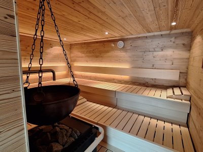 In der Sauna sitzen Personen auf Bänken aus Holz. Der Saunaofen steht in der Mitte. An der Decke sind Lampen.