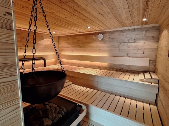 Finnische Sauna mit Aufgußautomat In der Sauna sitzen Personen auf Bänken aus Holz. Der Saunaofen steht in der Mitte. An der Decke sind Lampen.