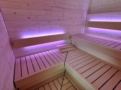 In einem Saunaraum sind die Wände aus Holz. Bunte Lichter sind sichtbar. Bänke aus Holz sind bereit für die Nutzung.