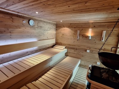 In einem Raum mit Holzvertäfelung stehen Bänke aus Holz. Im Hintergrund gibt es eine Sauna mit passenden Utensilien.