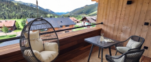 Auf dem Balkon stehen ein Tisch und zwei Stühle. Ein Hängesessel ist bereit zum Entspannen. Die Berge sind im Hintergrund sichtbar.