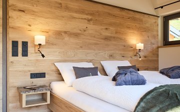 Zwei getrennte Betten stehen in einem Raum mit Holzwand. Es gibt Lampen und eine kleine Ablage.