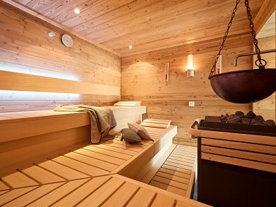 In einem Raum mit Holzwänden stehen Bänke aus Holz. Eine große Schüssel hängt über einem heißen Ofen mit Steinen.