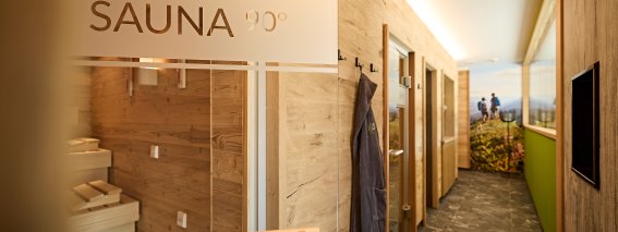Der Raum zeigt einen Saunabereich mit Holzregalen und offenen Türen. Menschen entspannen in der Nähe des Fensters.