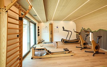In einem Raum gibt es Fitnessgeräte wie Laufband, Rudergerät und Fahrräder. Das Licht ist hell und der Boden ist aus Holz.