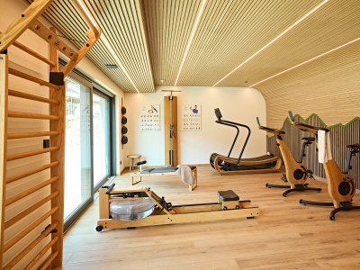 In einem Raum gibt es Fitnessgeräte wie Laufband, Rudergerät und Fahrräder. Das Licht ist hell und der Boden ist aus Holz.